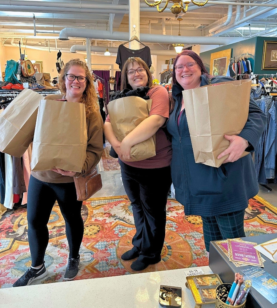 Three women holding brown paper bags in a store setting with colorful carpet and clothing racks.